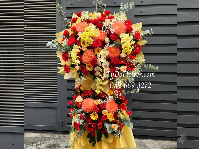 KCM0125167 KỆ HOA CHÚC MỪNG NGÀY THẦY THUỐC VN Thiết kế tông màu đỏ vàng cam, chủ đạo Hoa Cúc Mẫu Đơn Cam, Hoa Hồng Đỏ Ecuador, Lan Hồ Điệp Vàng