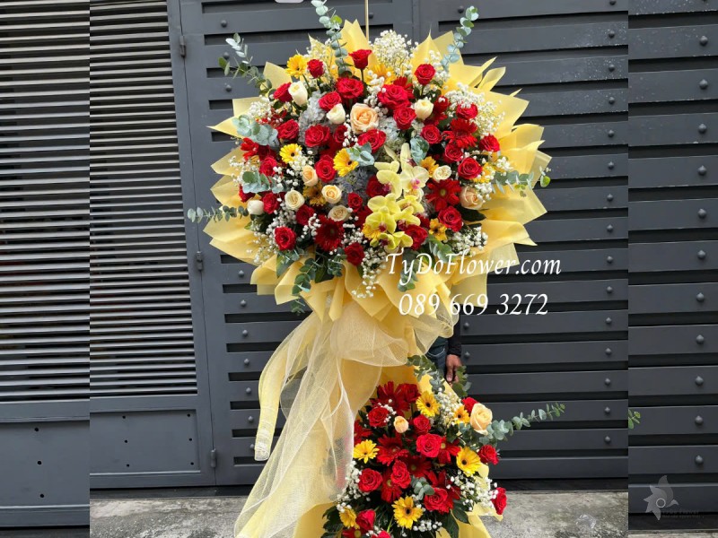 KCM0125172 KỆ HOA CHÚC MỪNG NGÀY THẦY THUỐC VN Thiết kế tông màu đỏ vàng cam, chủ đạo Hoa Hồng Đỏ Ecuador, Hoa Hồng Cam, Lan Hồ Điệp Vàng, Đồng Tiền