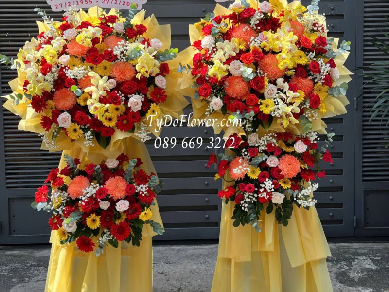 KCM0125174 ĐẶT TỪ 02 KỆ HOA CHÚC MỪNG NGÀY THẦY THUỐC VN Thiết kế tông màu đỏ vàng cam, chủ đạo Hoa Cúc Mẫu Đơn Cam, Hoa Hồng Đỏ Ecuador, Lan Hồ Điệp Vàng