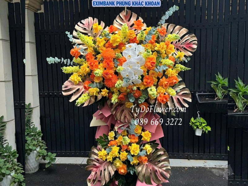 KCM0825620 KỆ HOA CHÚC MỪNG Thiết kế gam màu vàng cam, chủ đạo hoa hồng, lan hồ điệp, lá trầu bà nam mỹ