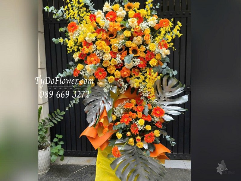 KCM0126028 KỆ HOA CHÚC MỪNG NGÀY THÀNH LẬP Thiết kế tông màu vàng cam, hoa hồng, hoa đồng tiền, lan vũ nữ, hoa hướng dương, lá trầu bà nam mỹ