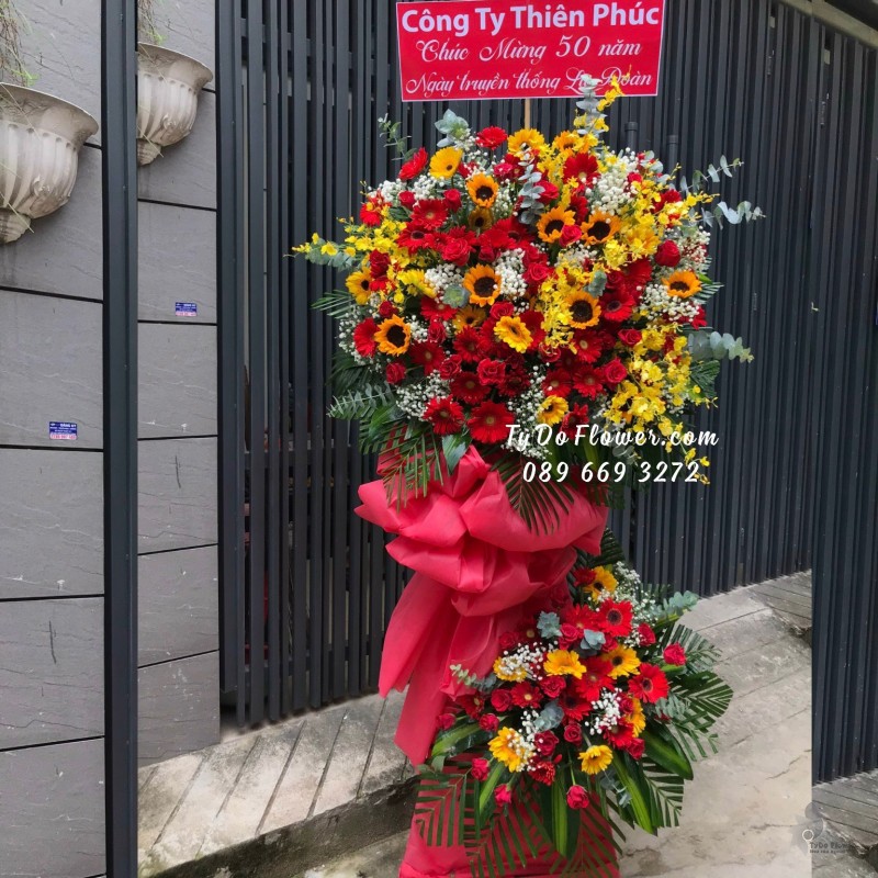 KCM09231042 KỆ HOA CHÚC MỪNG 50 NĂM NGÀY TRUYỀN THỐNG thiết kế Hoa Hồng Đỏ, Hoa Hướng Dương, Hoa Lan Vũ Nữ, Hoa Đồng Tiền Đỏ Vàng mix Baby Trắng, Lá Bạc