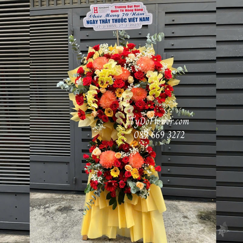 KCM0125167 KỆ HOA CHÚC MỪNG NGÀY THẦY THUỐC VN Thiết kế tông màu đỏ vàng cam, chủ đạo Hoa Cúc Mẫu Đơn Cam, Hoa Hồng Đỏ Ecuador, Lan Hồ Điệp Vàng