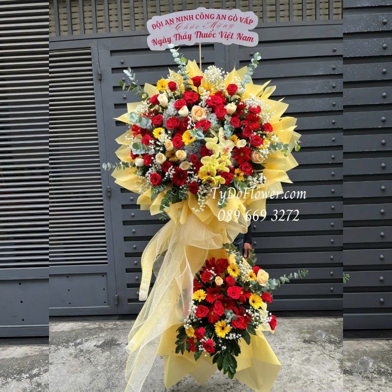 KCM0125172 KỆ HOA CHÚC MỪNG NGÀY THẦY THUỐC VN Thiết kế tông màu đỏ vàng cam, chủ đạo Hoa Hồng Đỏ Ecuador, Hoa Hồng Cam, Lan Hồ Điệp Vàng, Đồng Tiền