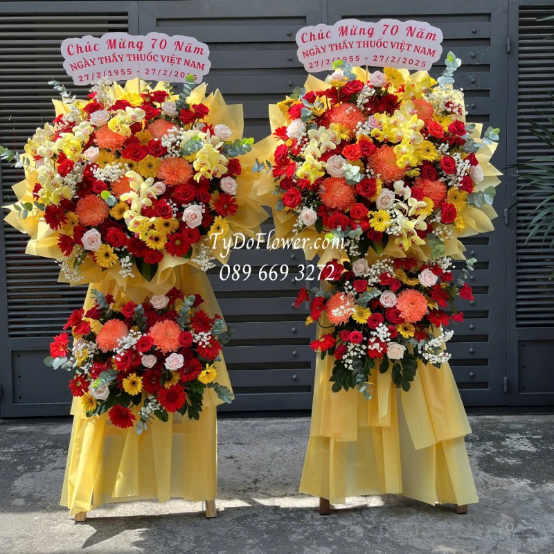 KCM0125174 ĐẶT TỪ 02 KỆ HOA CHÚC MỪNG NGÀY THẦY THUỐC VN Thiết kế tông màu đỏ vàng cam, chủ đạo Hoa Cúc Mẫu Đơn Cam, Hoa Hồng Đỏ Ecuador, Lan Hồ Điệp Vàng