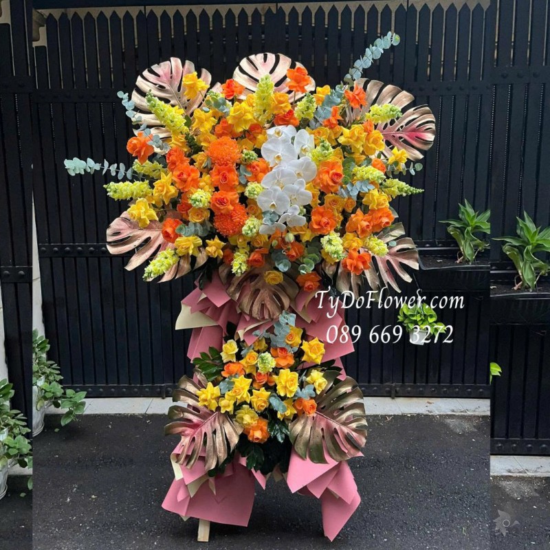 KCM0825612 KỆ HOA CHÚC MỪNG Thiết kế gam màu vàng cam, chủ đạo hoa hồng, lan hồ điệp, lá trầu bà nam mỹ