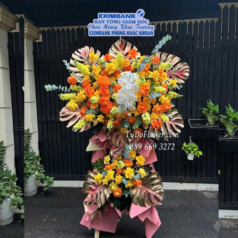 KCM0825620 KỆ HOA CHÚC MỪNG Thiết kế gam màu vàng cam, chủ đạo hoa hồng, lan hồ điệp, lá trầu bà nam mỹ