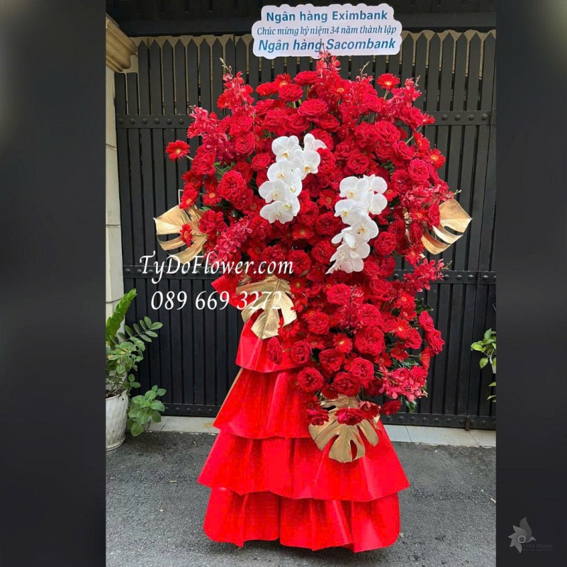 KCM0126027 KỆ HOA CHÚC MỪNG KỶ NIỆM NGÀY THÀNH LẬP Thiết kế hoa đồng tiền đỏ Red Ohara Rose, Lan Hồ Điệp, Lá Trầu Bà Nam Mỹ, Lan Mokara, Đồng Tiền Đỏ